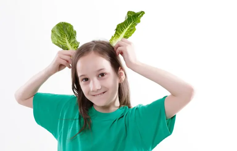 Young girl wearing a green shirt holds two lettuce leaves above her head, smiling at the camera.