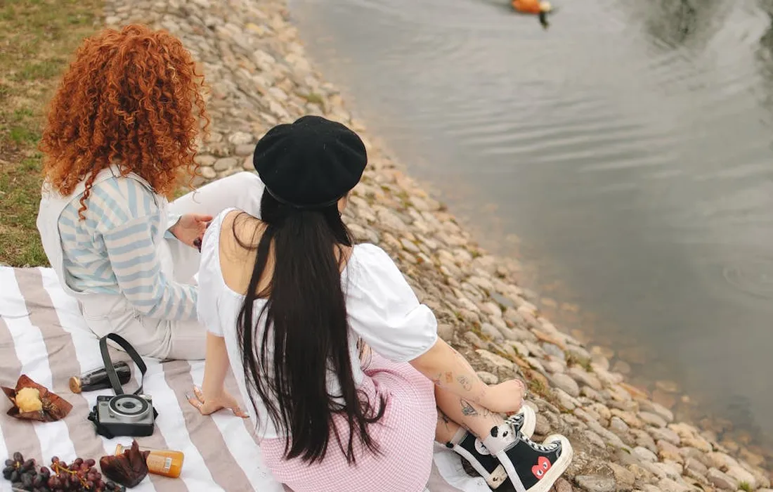 Two people sit on a striped blanket by a pond, watching a duck in a relaxed backyard setting.