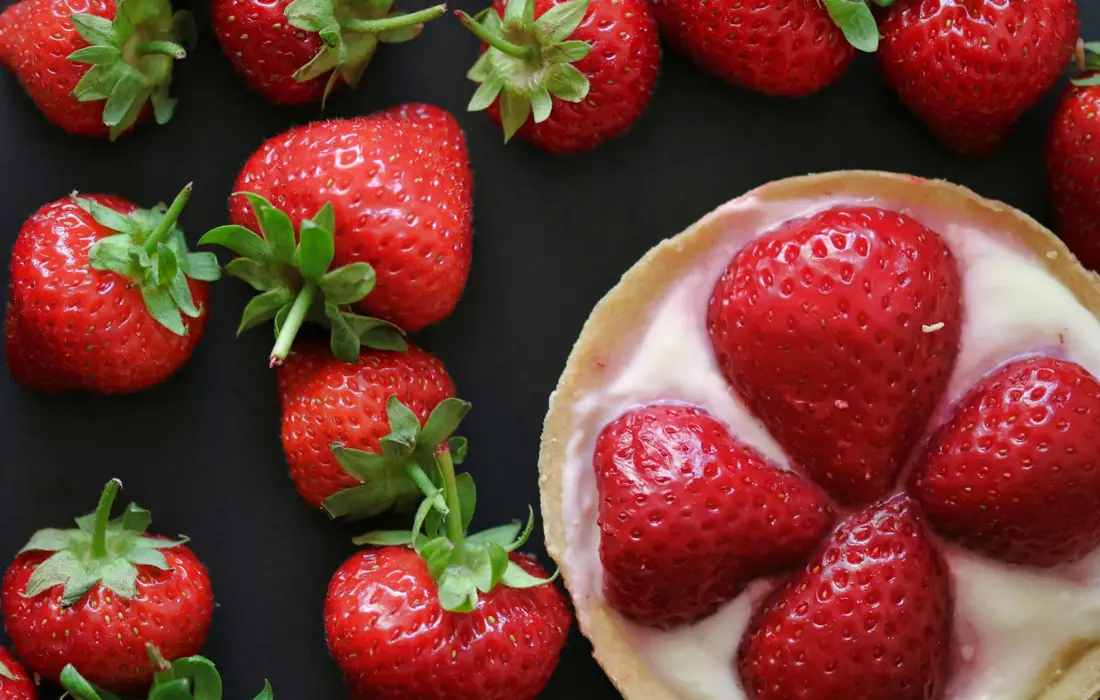 Close-up of fresh strawberries around a berry tart on a dark surface, illustrating seasonal berry treats from the farm.