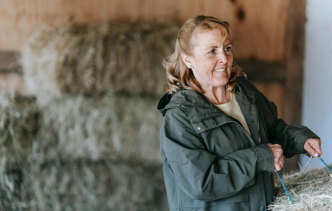 Smiling woman in a green jacket standing in a barn with stacked hay bales in the background, holding a rope.