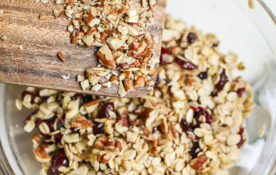 Close-up of a wooden spoon scooping chopped nuts and dried fruits over a bowl of granola, illustrating dried fruits such as raisins and cranberries used in livestock feed discussions.