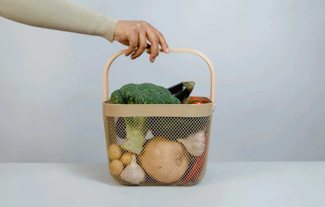 Person's hand lifting a beige mesh basket filled with broccoli and assorted vegetables against a neutral background.