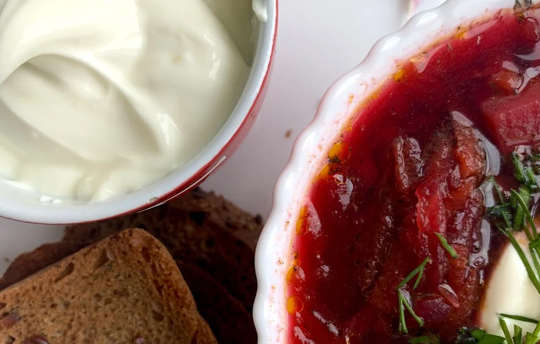 Beet soup (borscht) in a white dish with dill garnish, accompanied by a small bowl of sour cream and slices of dark bread.
