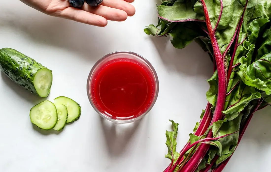 Beet greens with red stems and a glass of beet juice on a white surface, with cucumber slices nearby.