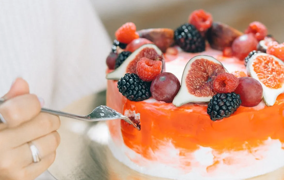 A person with a ring uses a cake server to cut into a berry-topped frosted cake, decorated with raspberries and blackberries.