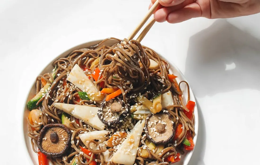 Bowl of mushroom and vegetable soba noodles with sesame seeds, being lifted with chopsticks