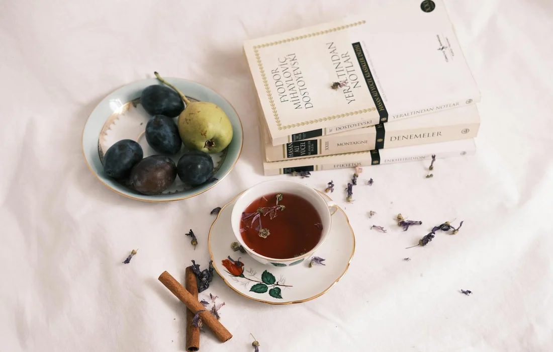 Flat lay of a white surface with a bowl of pears and grapes, a cup of tea on a saucer, cinnamon sticks, sprigs of lavender, and a small stack of books.
