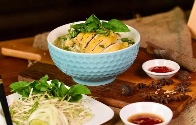 Blue textured bowl of pasta with sliced chicken and fresh herbs on a wooden table, with small bowls of sauce and a side salad nearby.