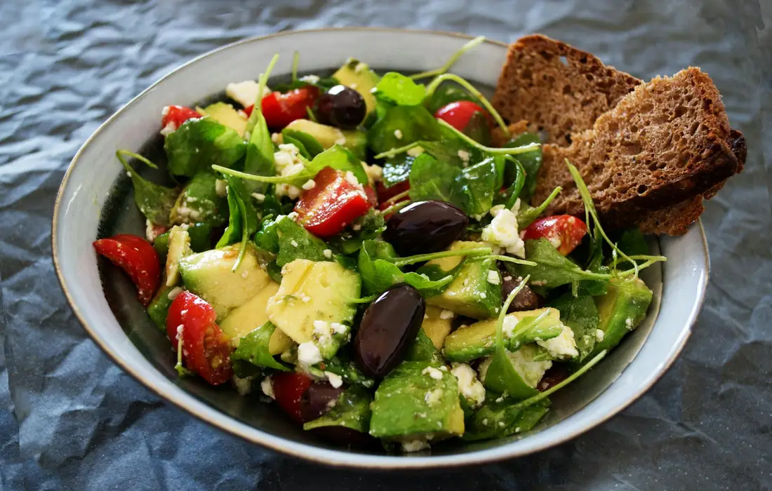 Bowl of mixed leafy greens with avocado, olives, red peppers, feta cheese, and slices of dark bread on the side.