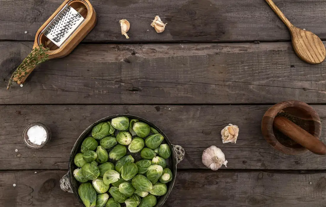 Top-down view of a bowl of Brussels sprouts on a rustic wooden table with garlic, a grater, and wooden spoon nearby