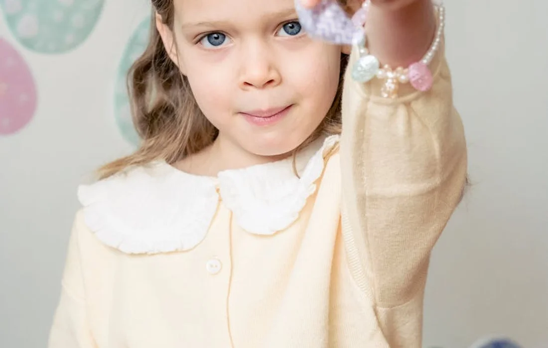 Young girl with light-colored clothing holding a string of pastel beads indoors.