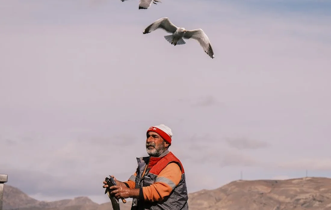 Man in an orange jacket and red beanie stands outdoors in a dry landscape, holding a device as several birds fly overhead.
