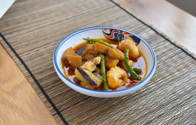 A bowl of mixed cooked vegetables in sauce on a woven placemat, featuring squash, eggplant, and green beans.