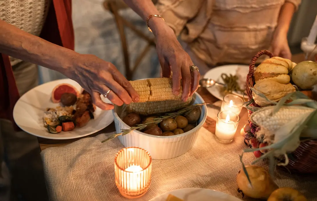 Hands handling a corn cob over a bowl of corn on a dining table, with plates and candles nearby, illustrating food handling context for corn safety in pig feed.