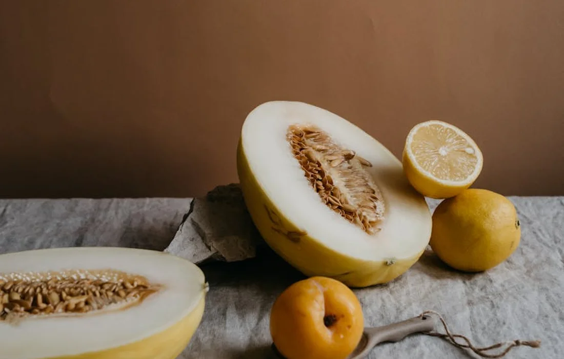 Halved cantaloupe with seeds exposed on a table, accompanied by a lemon and small yellow fruits