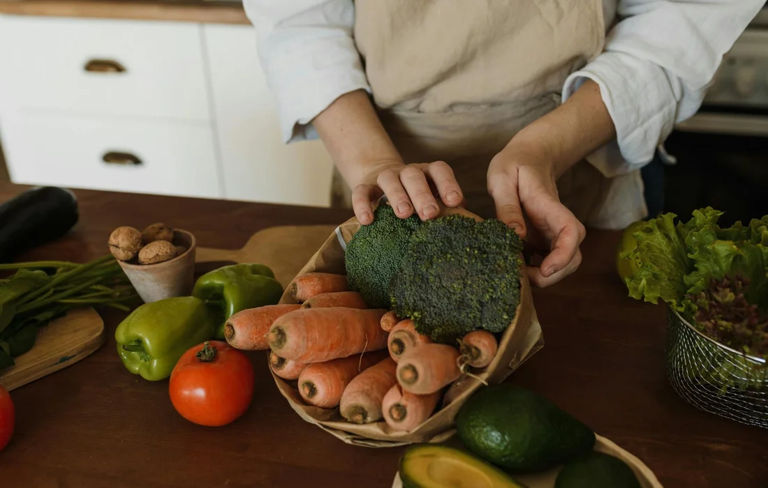 A person in a kitchen apron holds a broccoli head over a basket of baby carrots, with peppers and greens visible on the counter.