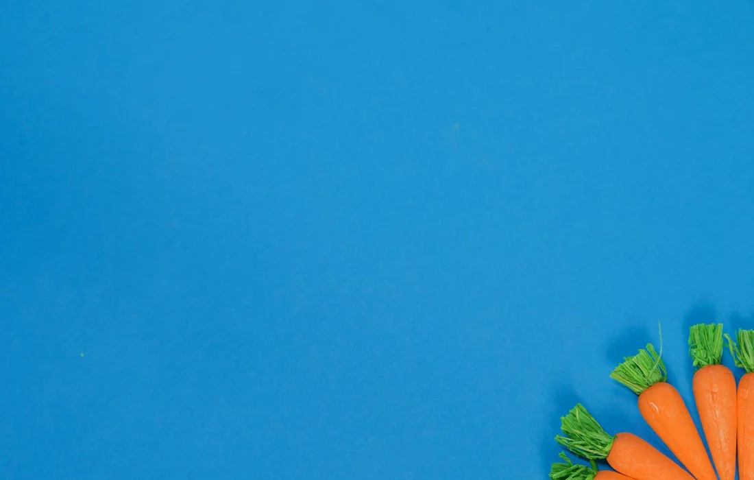 Fresh carrots with green tops arranged in the bottom right corner against a bright blue background.
