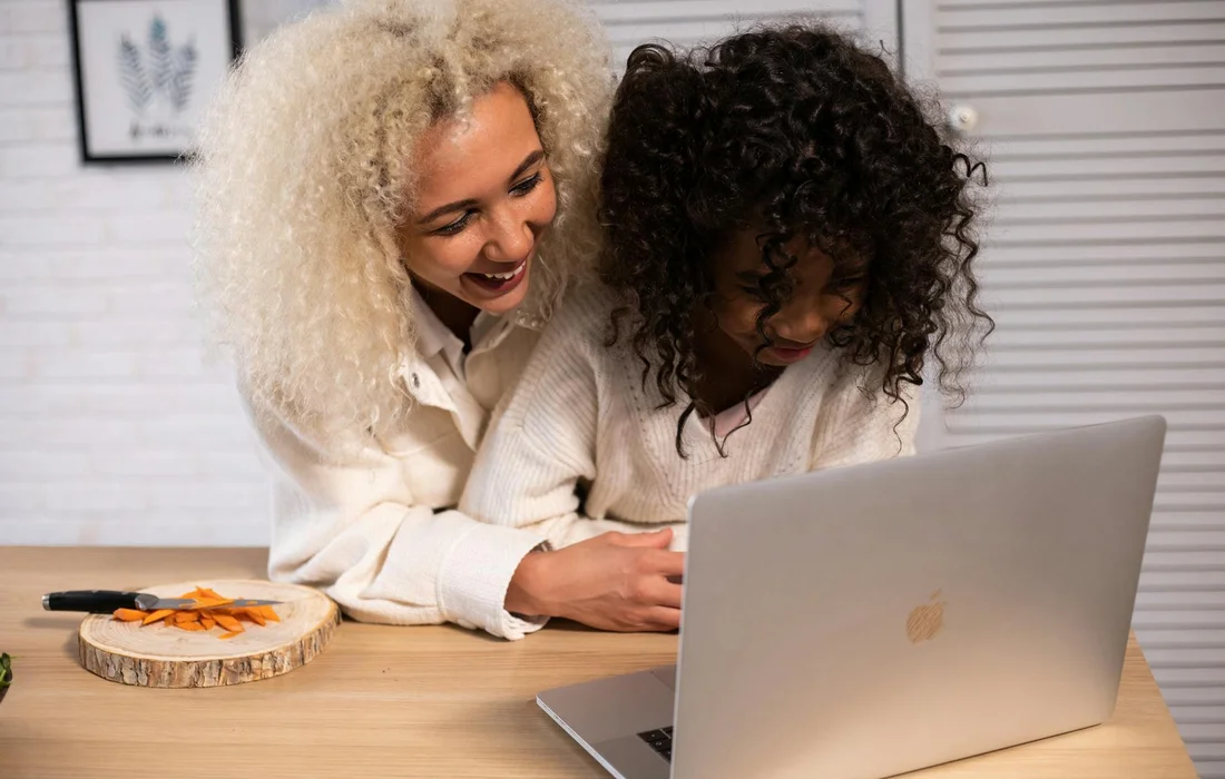 Two women looking at a laptop on a kitchen table with a small plate of carrot slices nearby, illustrating a discussion about garden treats for chickens.