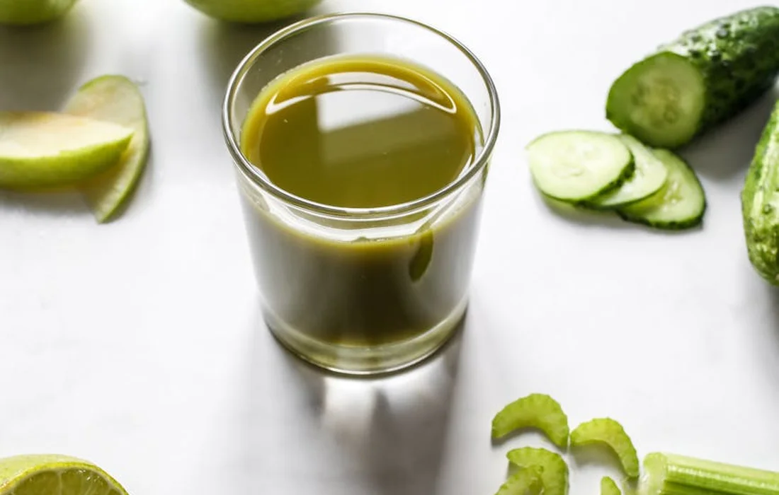 Glass of green juice surrounded by celery stalks and cucumber slices on a white surface, illustrating fresh vegetables that can complement a rabbit’s diet.
