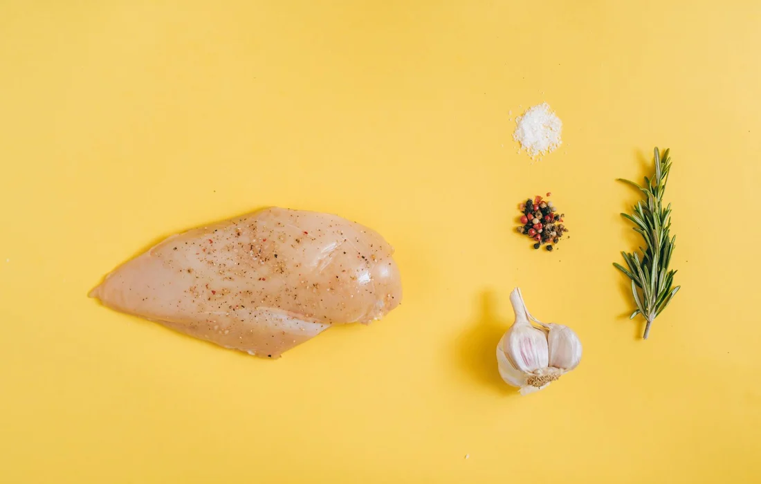 Raw chicken breast with garlic, a rosemary sprig, pepper, and coarse salt on a bright yellow background.