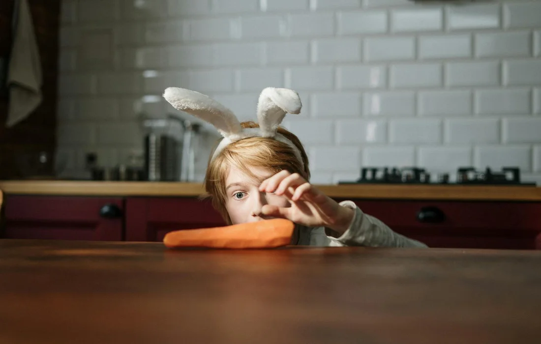 Child wearing bunny ears reaches for a carrot on a kitchen table.