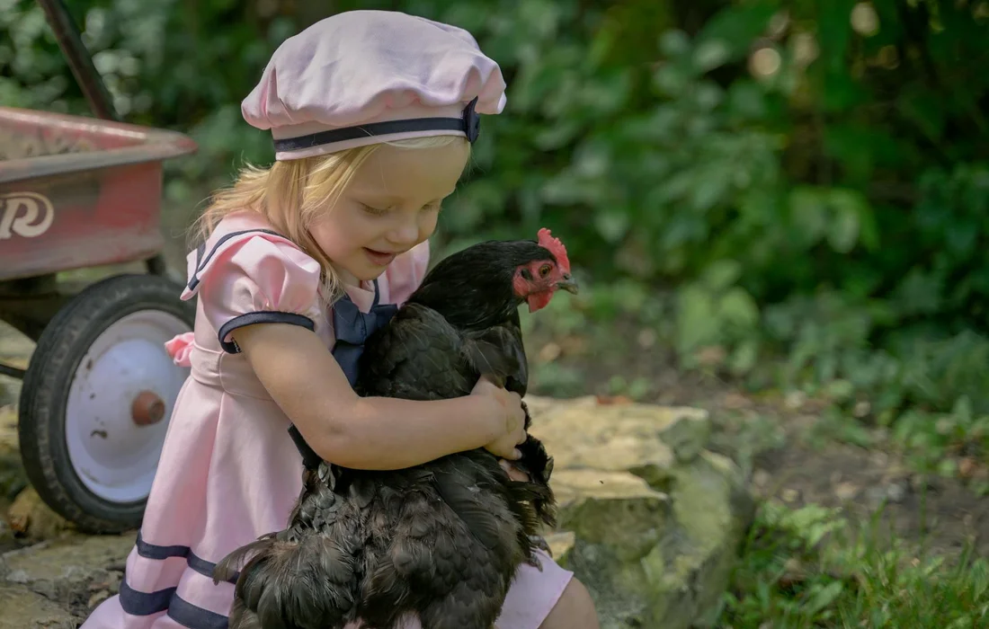 A young girl wearing a pink dress and pink hat hugs a black chicken in a sunny, green garden.