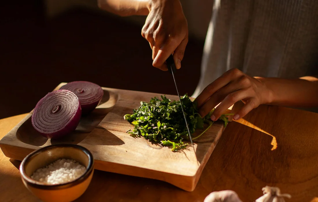 Close-up of hands chopping fresh cilantro on a wooden cutting board, with a sliced red onion and a small bowl nearby.