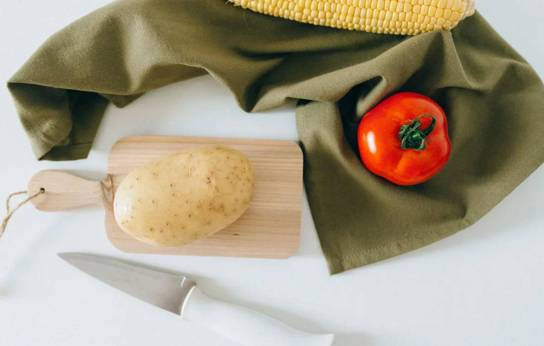 Corn on the cob, a potato on a cutting board, and a red tomato on a green cloth, with a knife nearby