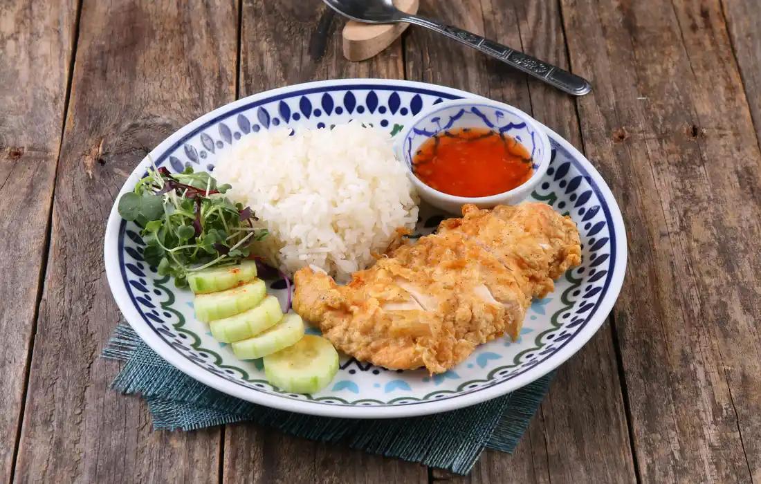 Plate with rice, fried chicken, cucumber slices, and dipping sauce on a wooden table, viewed from above.