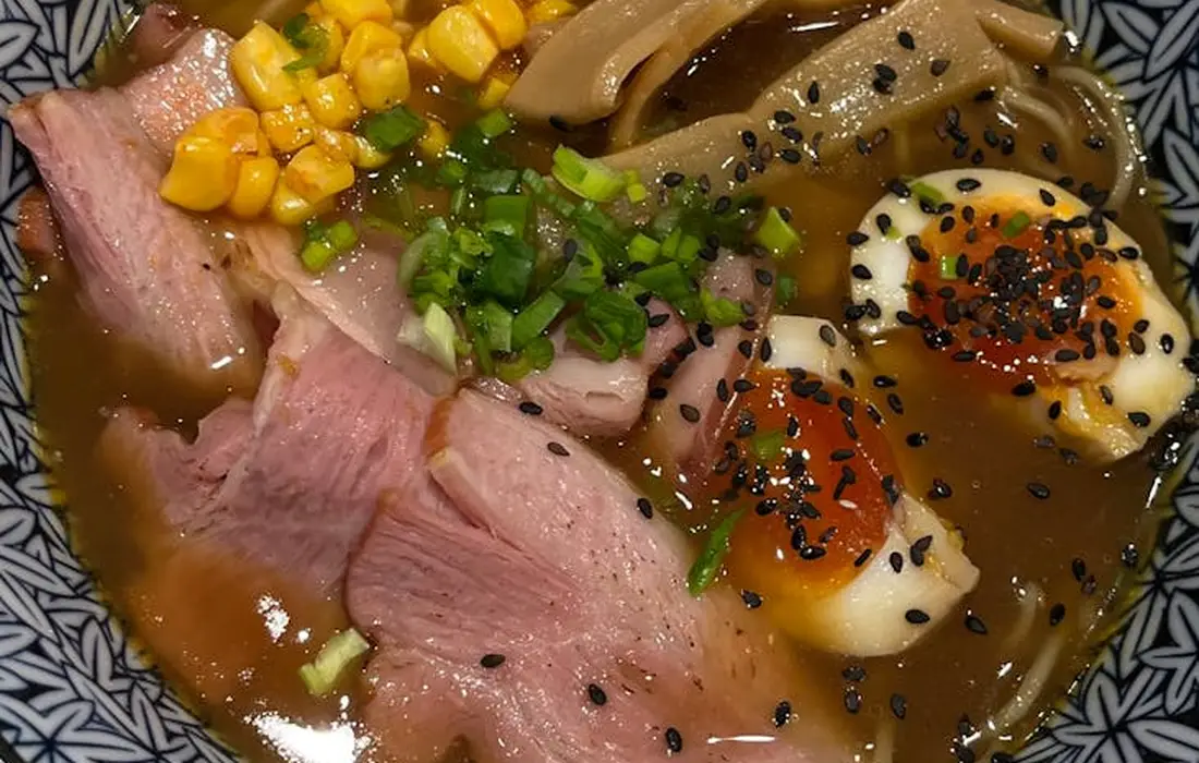 A bowl of ramen with slices of pork, corn, chopped green onions, and halved soft-boiled eggs topped with sesame seeds.