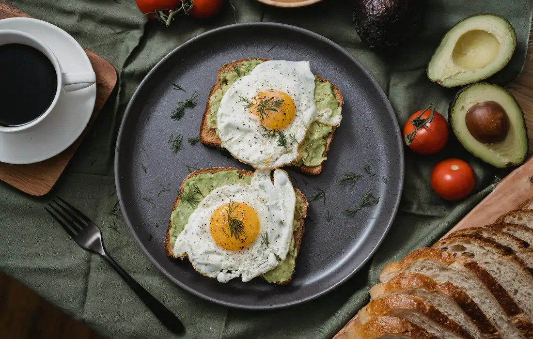 Two sunny-side-up eggs on avocado toast with herbs on a dark plate, surrounded by tomatoes, avocado halves, bread, and a cup of coffee on a green tablecloth.