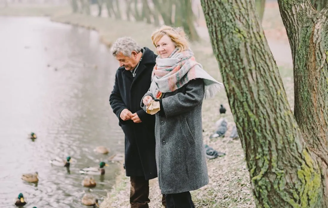 Couple feeding ducks by a lakeside with trees along the shore.