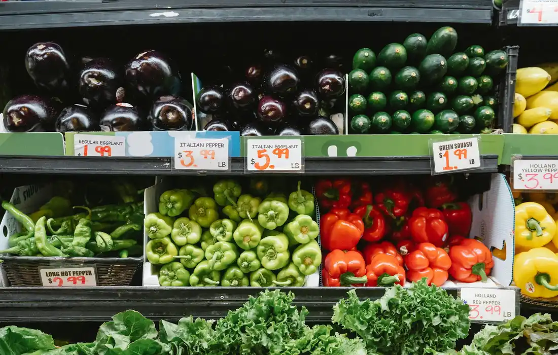 Market display with purple eggplants, green peppers, red bell peppers, and leafy greens.