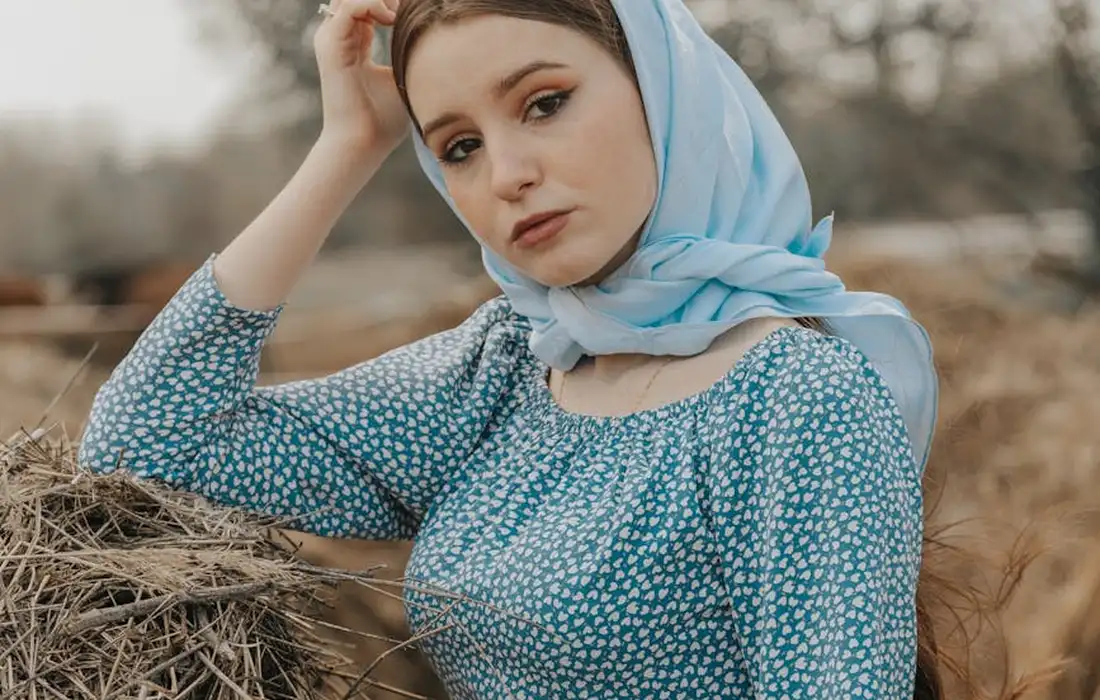 A woman wearing a blue floral dress and a light blue scarf stands beside a hay bale on a farm.