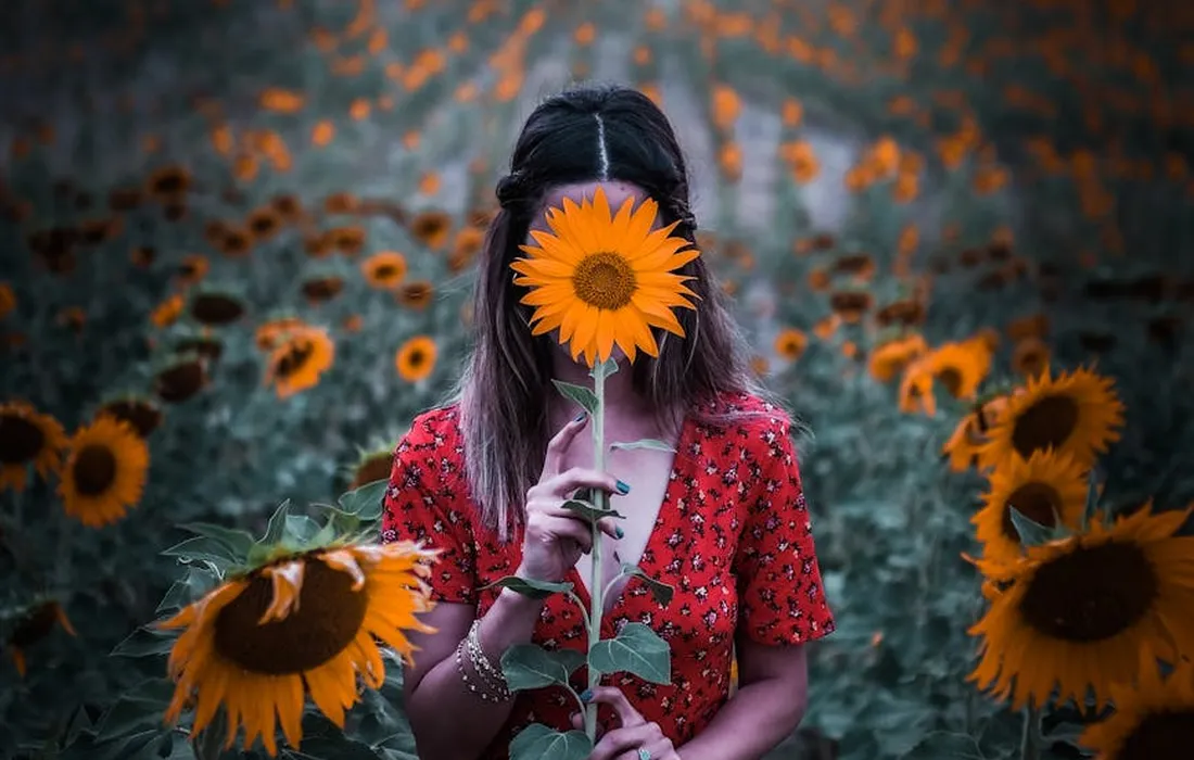 A person with shoulder-length dark hair wearing a red dress stands in a field of sunflowers, holding a sunflower up to their face.