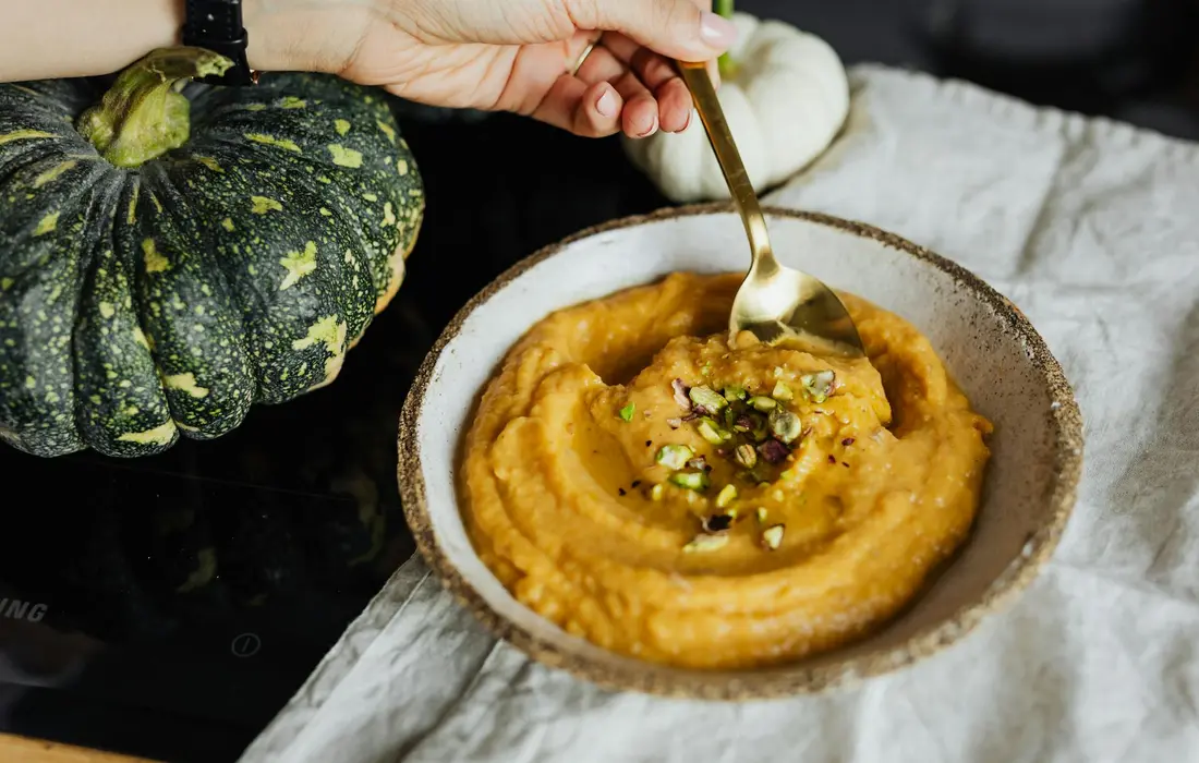 A bowl of creamy orange squash puree with a spoon, surrounded by green squash gourds and a light cloth.