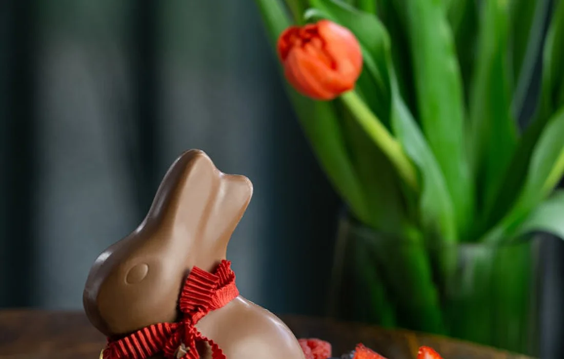 Chocolate bunny figurine with a red ribbon in front of a vase of tulips