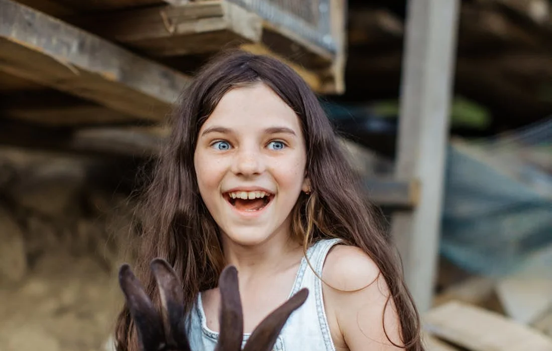 A young girl with long dark hair and a surprised expression holds a brown rabbit inside a rustic, barn-like space.