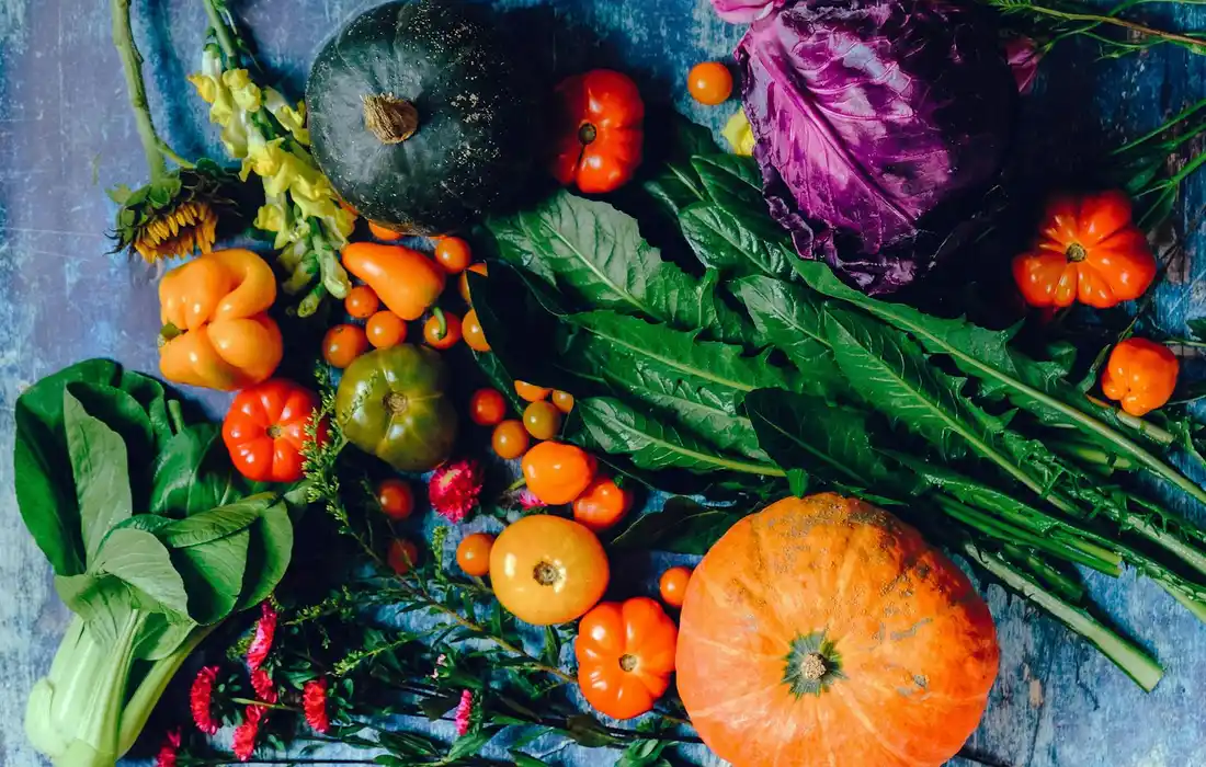 Assorted fresh vegetables including pumpkins, peppers, leafy greens, and squash arranged on a wooden surface.