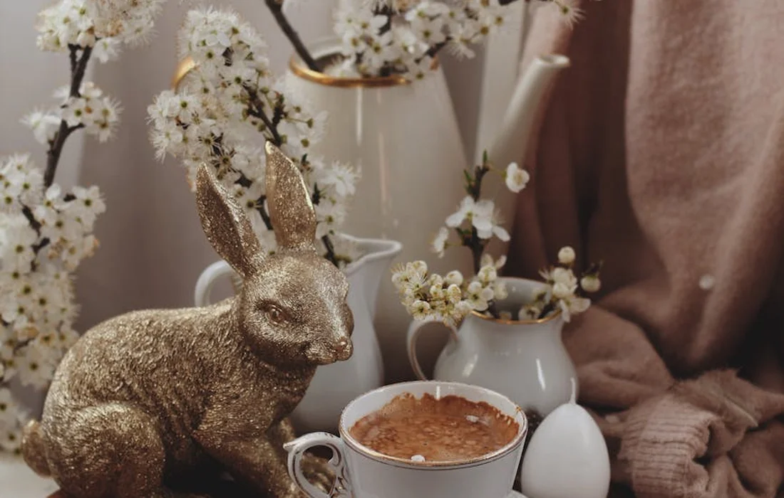 Glittery gold bunny statue beside a cup of cocoa and white blossoms on a soft beige background.