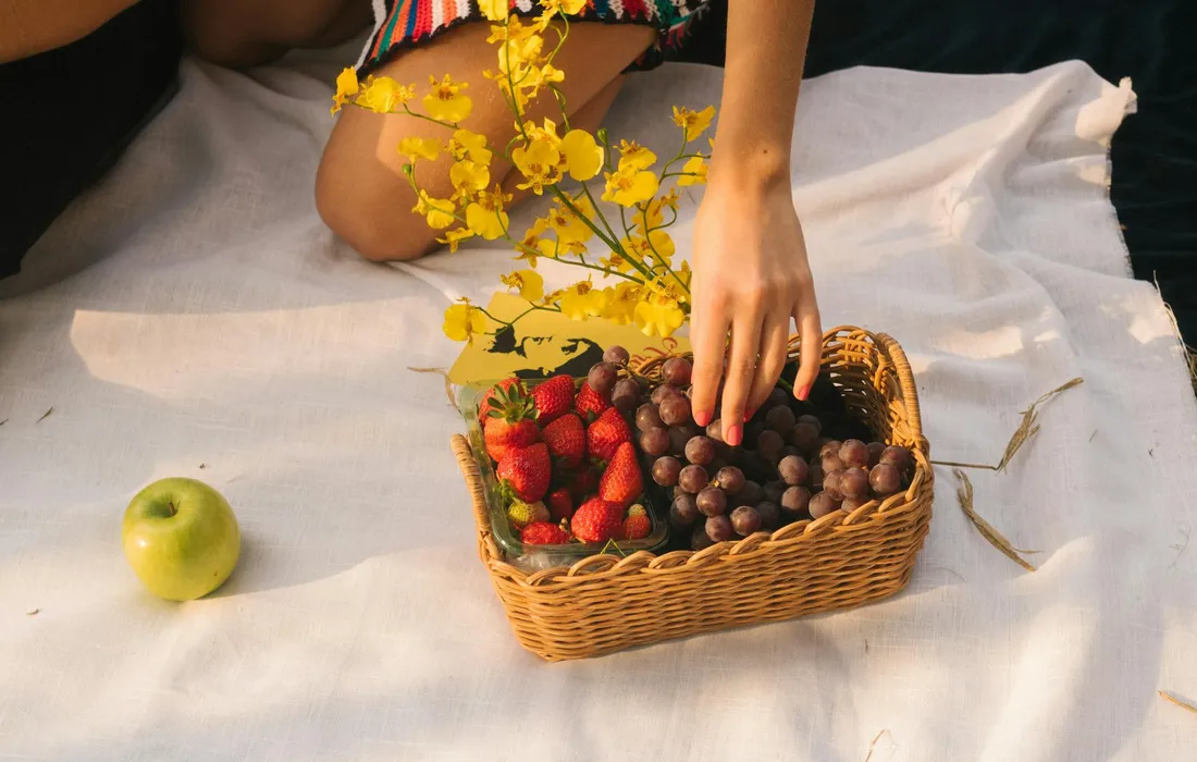 A hand reaching into a wicker basket filled with strawberries and grapes on a white blanket, with a green apple nearby and a small yellow flower arrangement in the background.