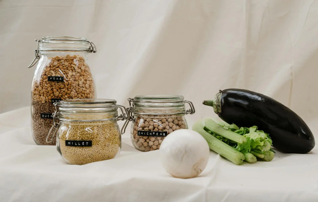 Still-life with celery stalks and a white onion beside glass jars of grains on a pale backdrop