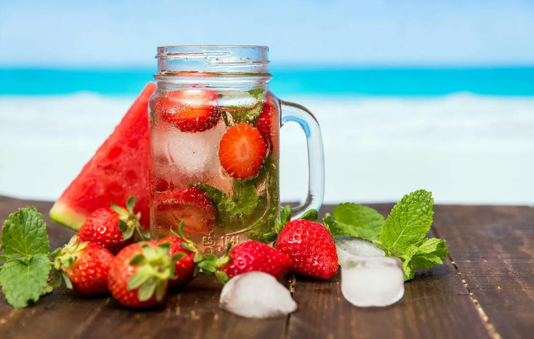 A mason jar filled with strawberry-infused water sits on a wooden table, surrounded by fresh strawberries, mint, and ice cubes with a beach scene in the background.