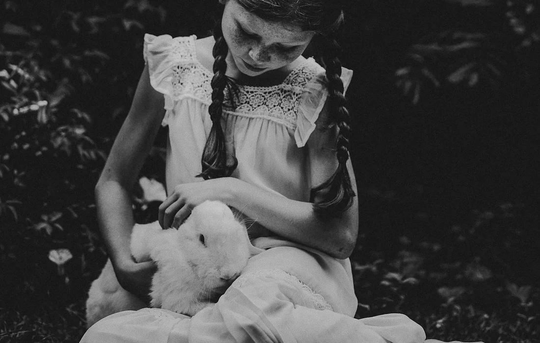 Black-and-white photo of a young girl with two braids wearing a white dress, sitting outdoors and gently cradling a white rabbit in her lap.