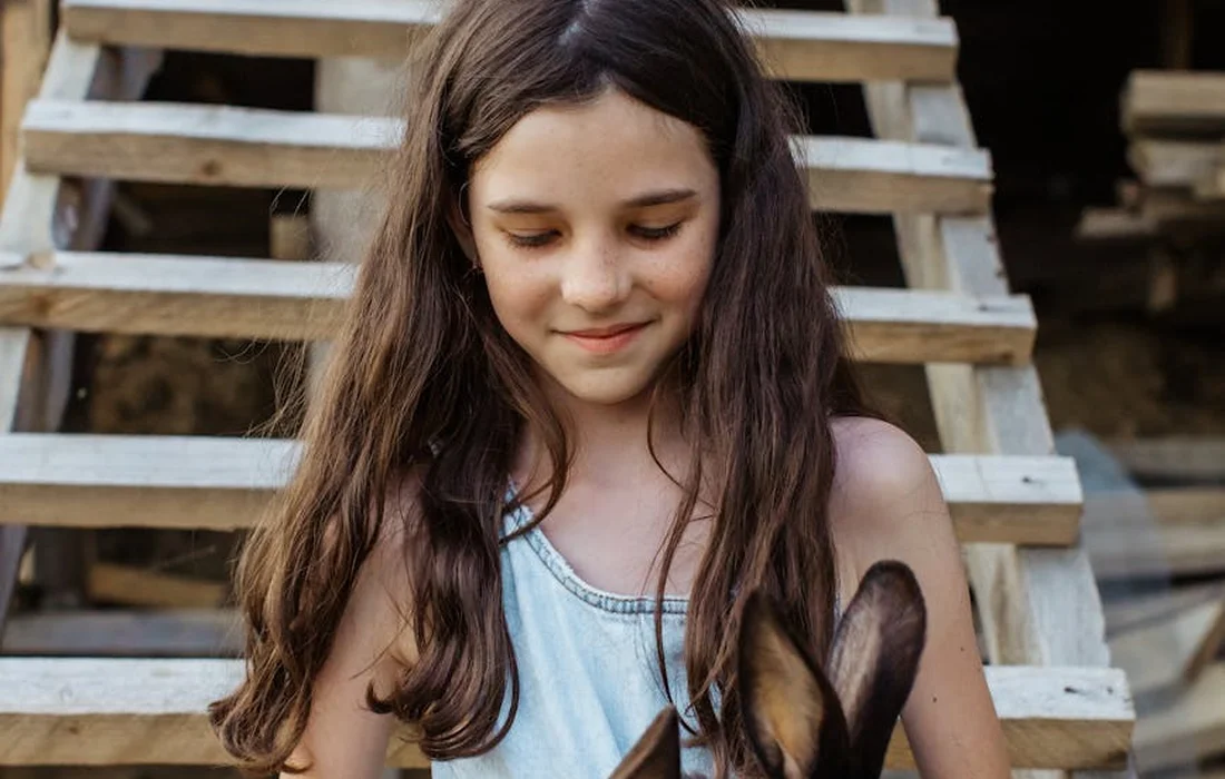 A young girl with long brown hair in a light blue top looks down at a rabbit she is holding, with wooden stairs in the background.