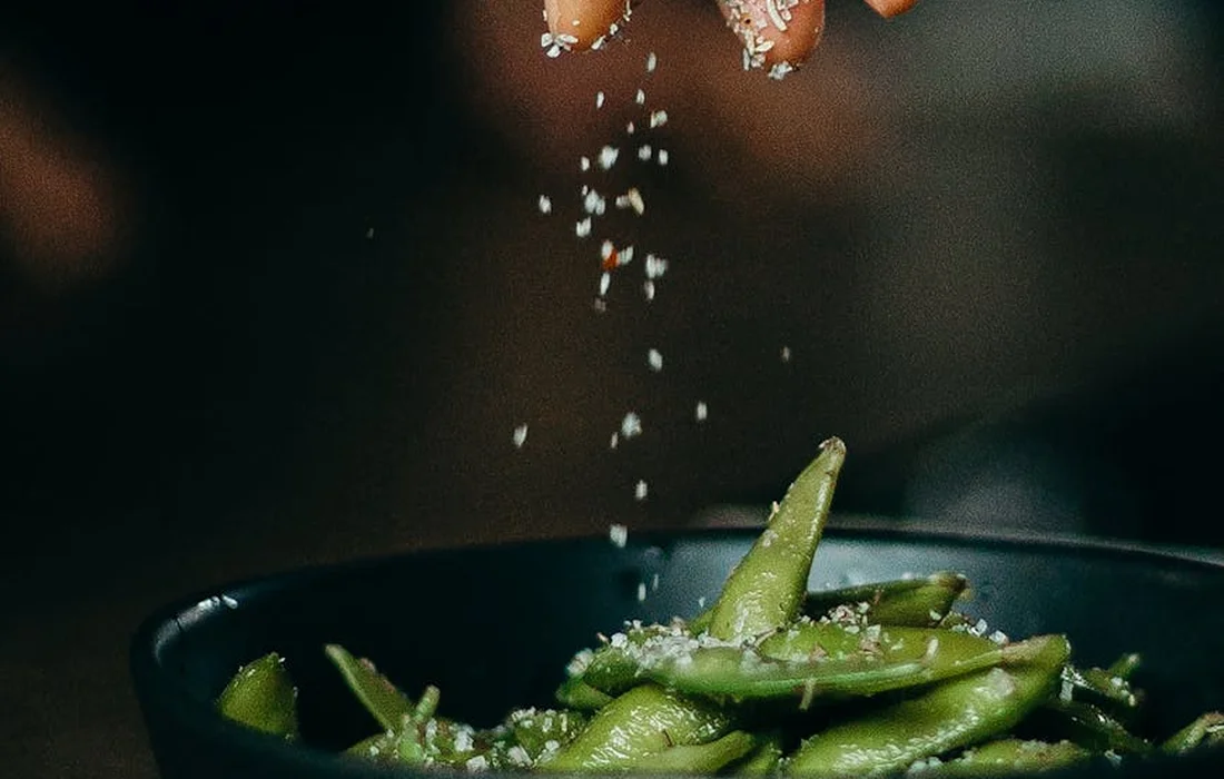 A hand sprinkles salt crystals over a bowl of fresh green beans, illustrating careful mineral and salt supplementation.