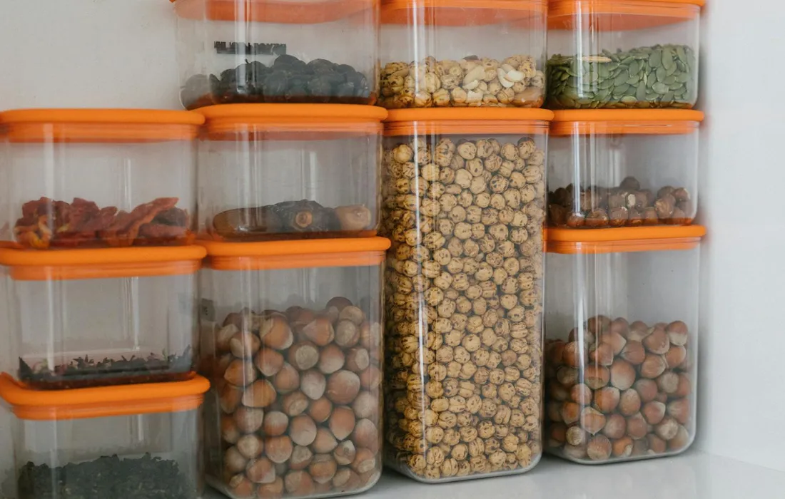 Clear plastic storage containers with orange lids filled with various grains and seeds, neatly stacked on a shelf