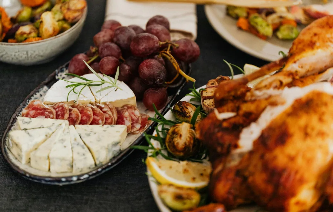 A rustic spread on a dark table featuring grapes on the stem, a cheese board with soft cheeses and slices of salami, fresh rosemary sprigs, lemon wedges, and a roasted chicken, illustrating foods that may be considered for poultry.