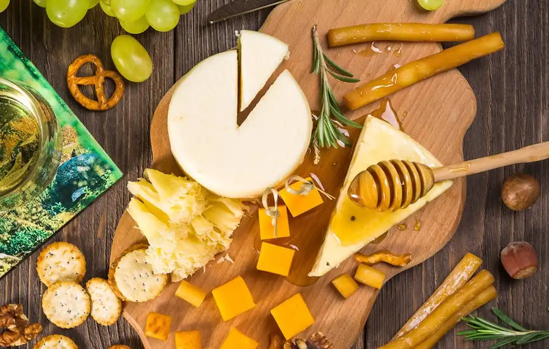 A cheese board with green grapes, assorted cheeses cut into cubes, a honey dipper, crackers, rosemary, and bread crisps arranged on a wooden surface.