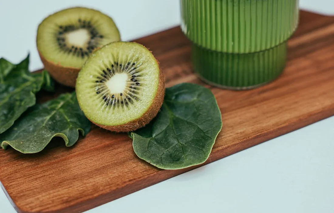 Kiwi halves and fresh leafy greens on a wooden board with a green cup in the background.